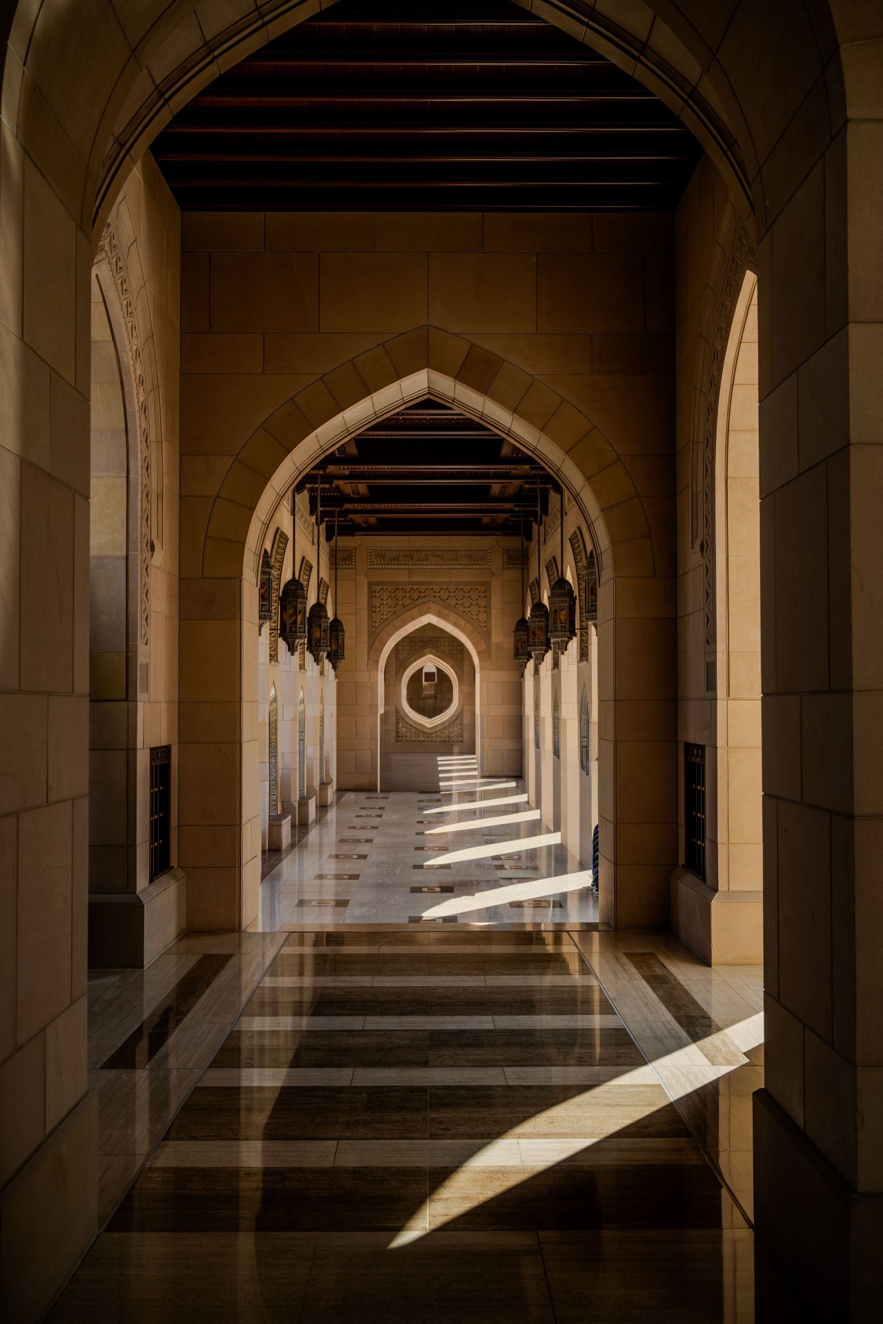Arched corridor of the Sultan Qaboos Grand Mosque in Muscat, warm sunlight falling across polished marble